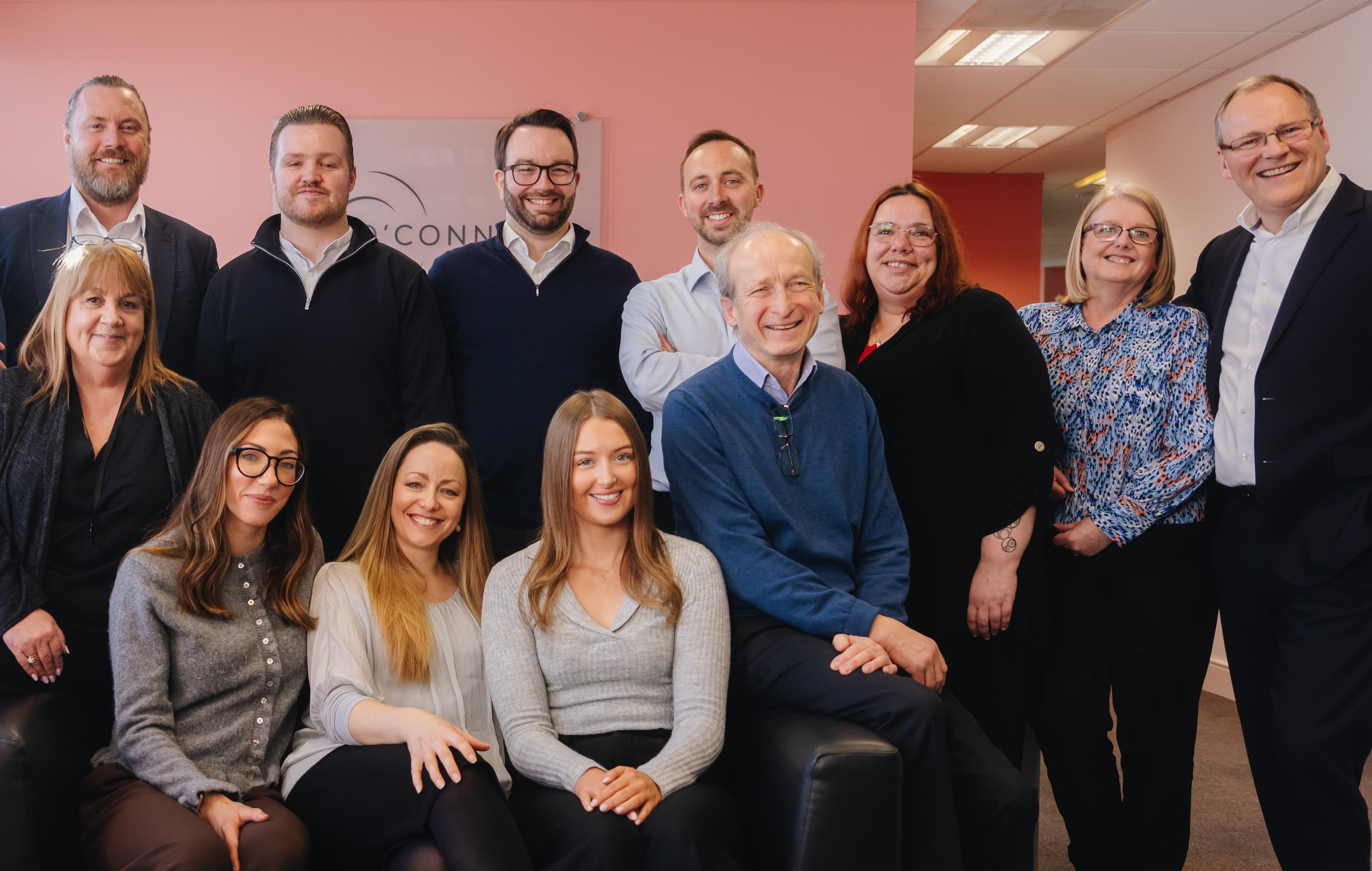 The team at O'Connors, one row is sat on a couch and the second row is standing.  The background is a blush coloured wall with the O'Connors logo visible.