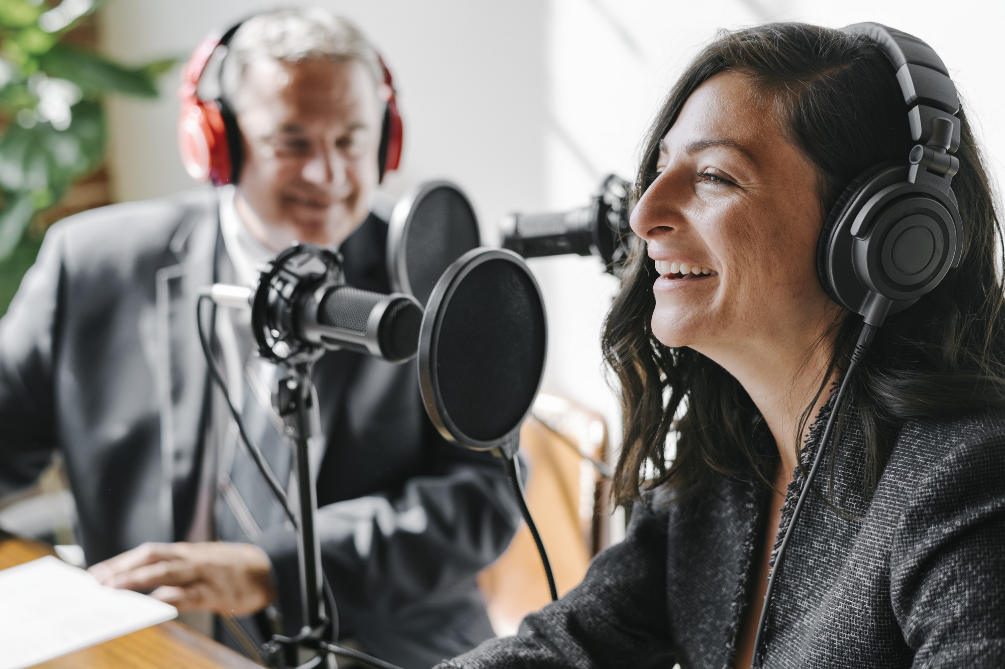 A smiling woman wearing black over ear headphones and a smiling man wearing red over ear headphones sitting in front of two microphones.