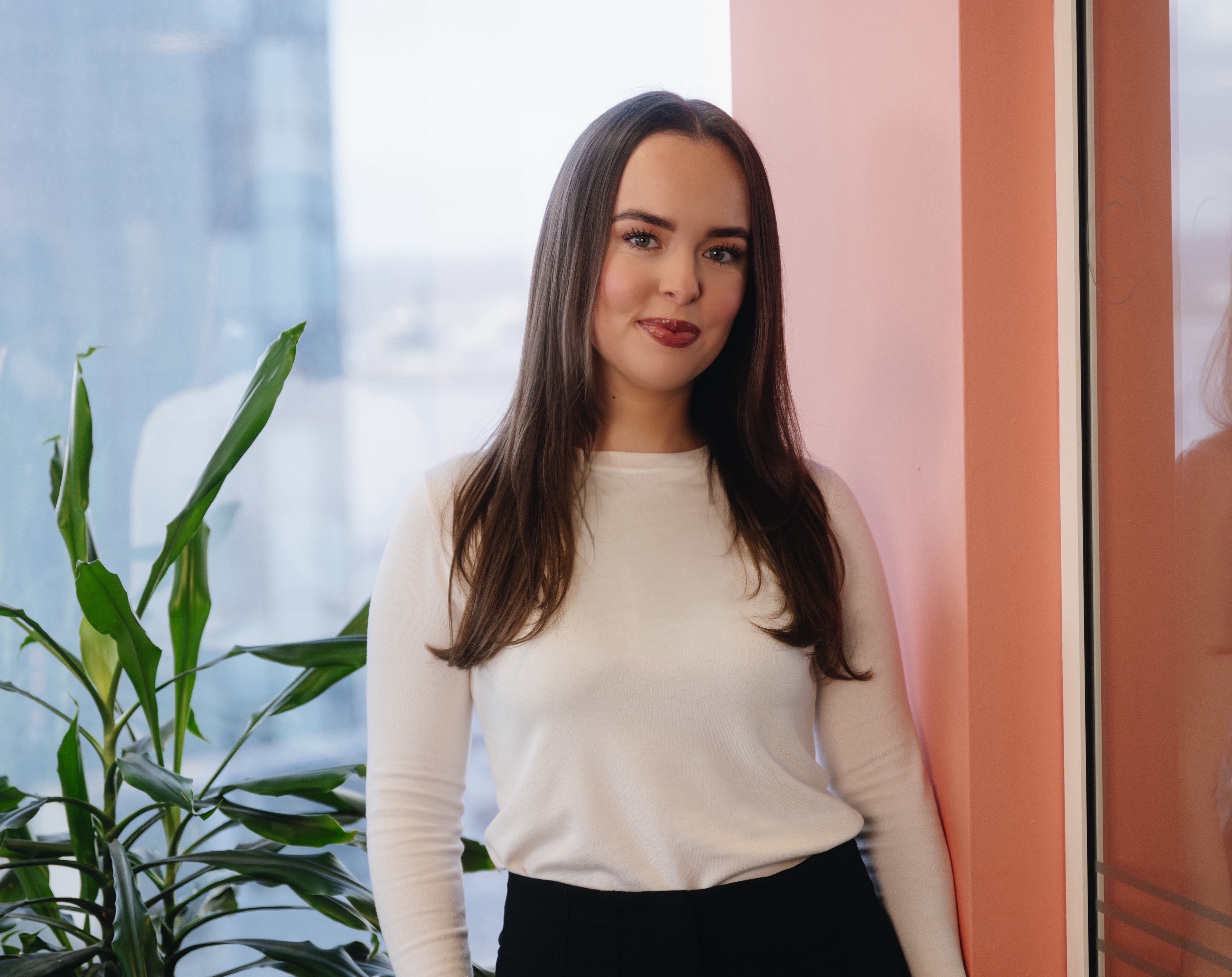 Mollie smiling.  She is standing in front of a window with a blush coloured pillar to the right and a tall green plant to the left.