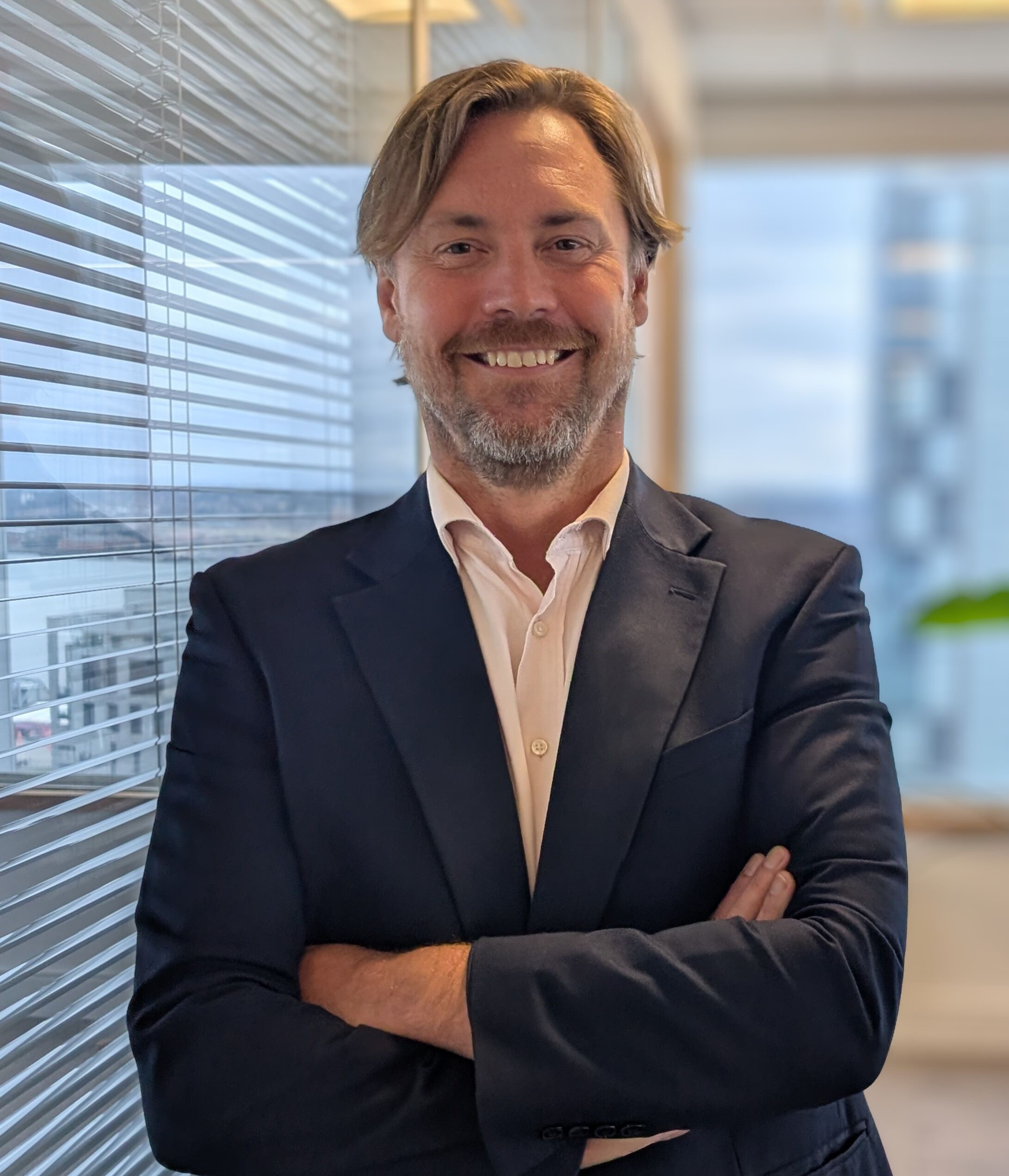 David smiling, arms crossed.  There is a glass partition with vertical blinds to his left and a window behind him with a green leaf visible.