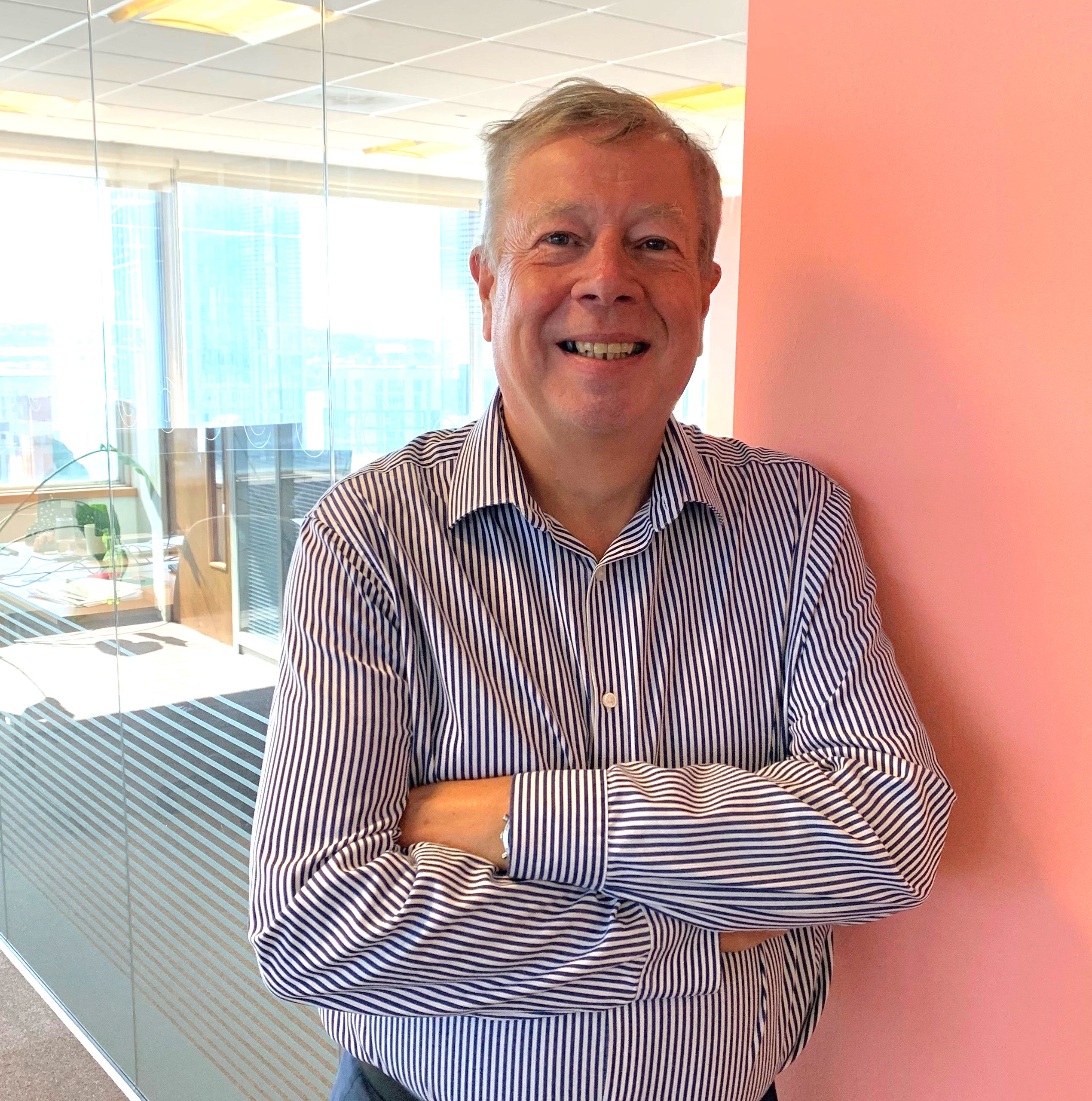 Laurence with his arms crossed and smiling.  He is leaning on a blush coloured wall with desks and a window in the background.