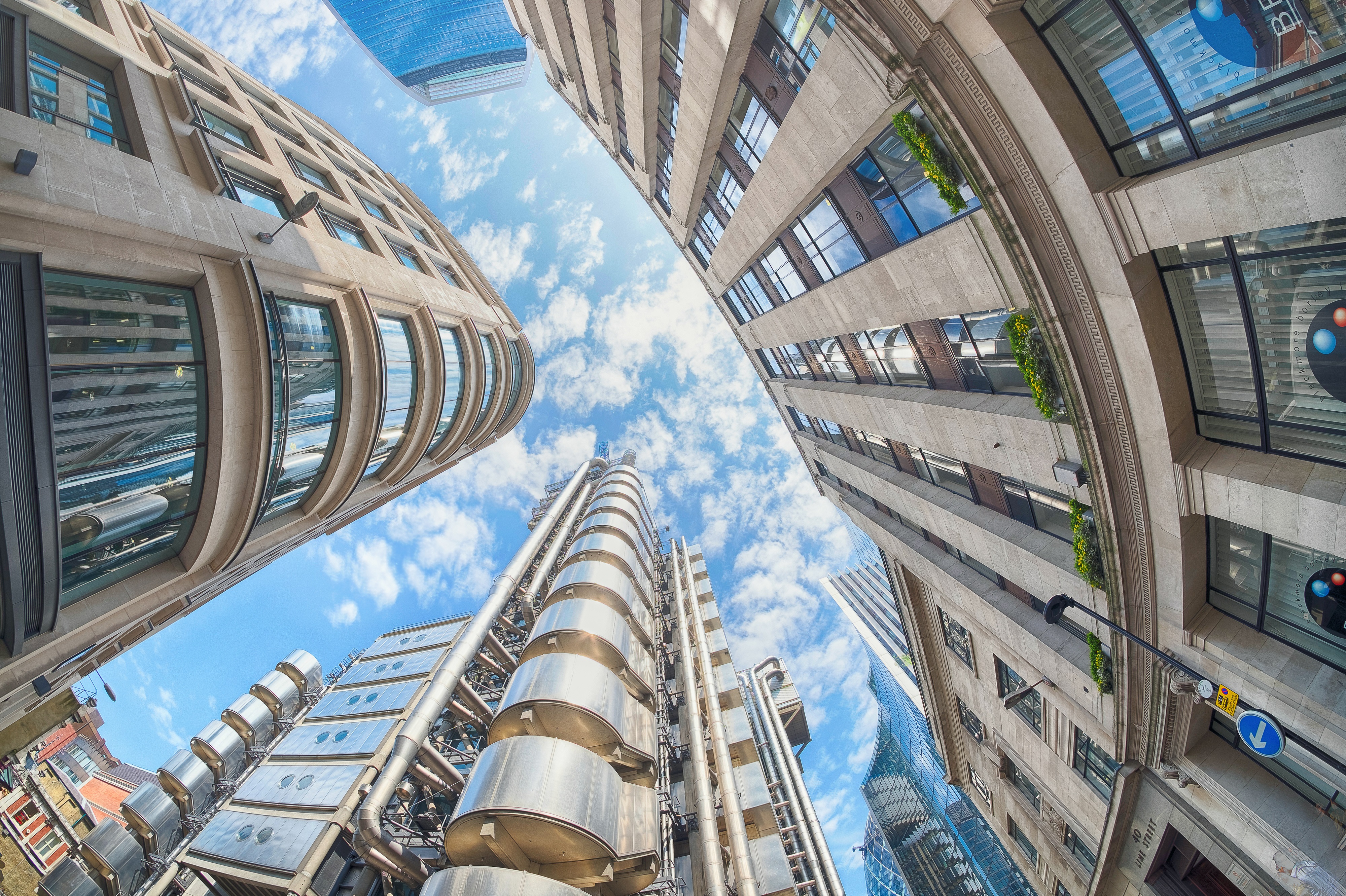 An upwards view of office blocks with a blue sky and white clouds above them.  The image is slightly curved.