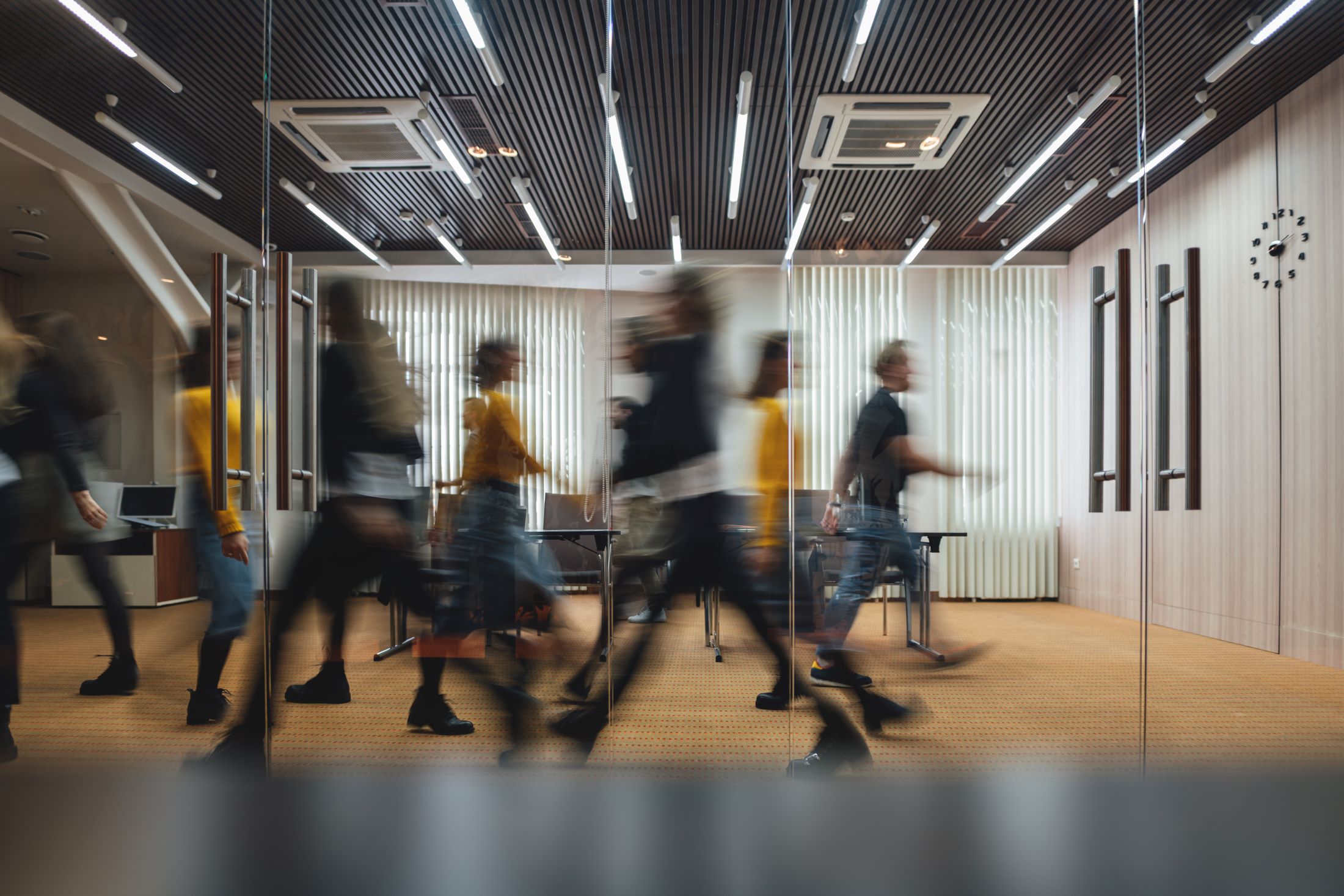 Blurred image of people walking around an office behind floor to ceiling glass doors.