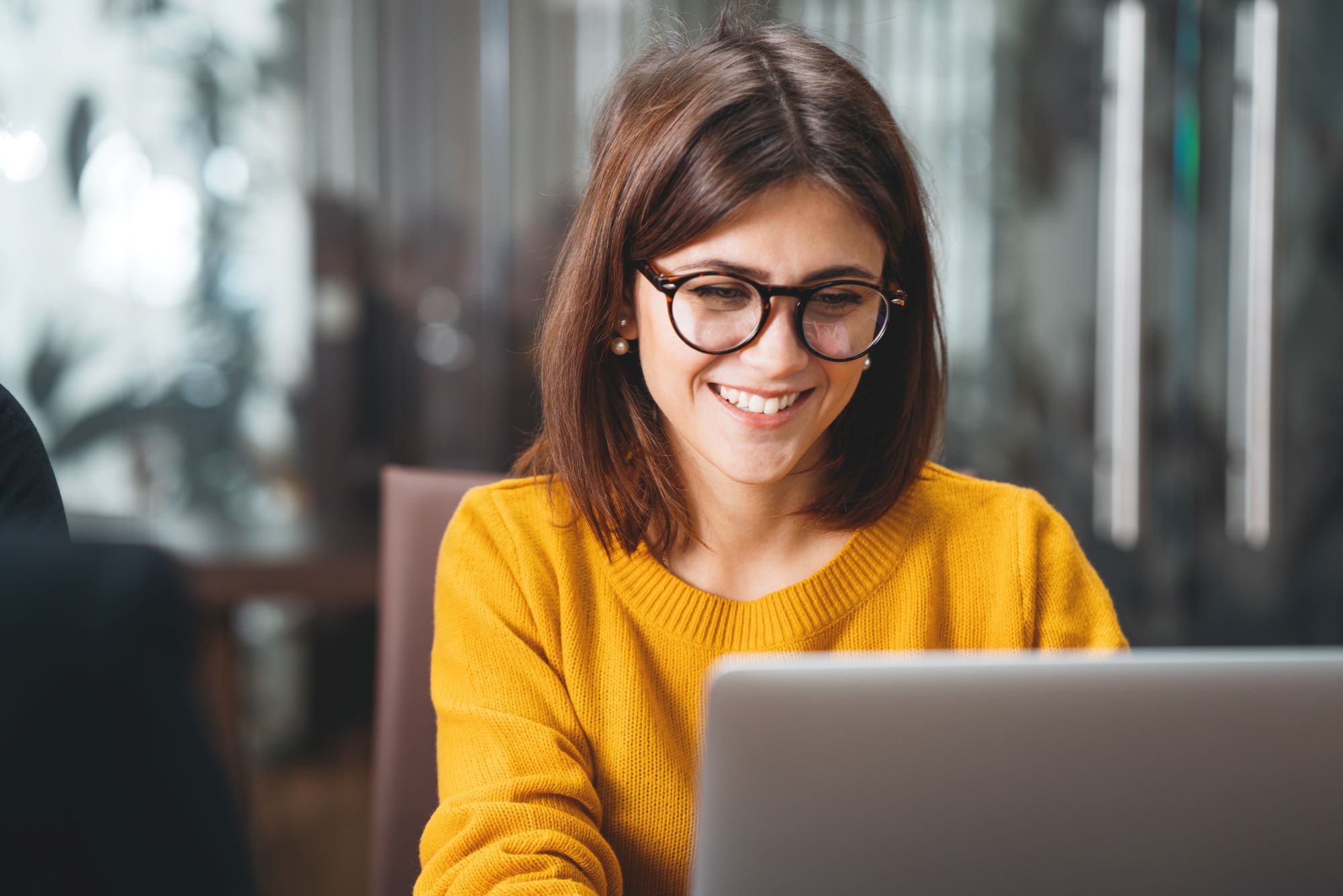 Smiling lady wearing glasses sat in front of a glass partition looking at her open laptop on the desk in front of her.