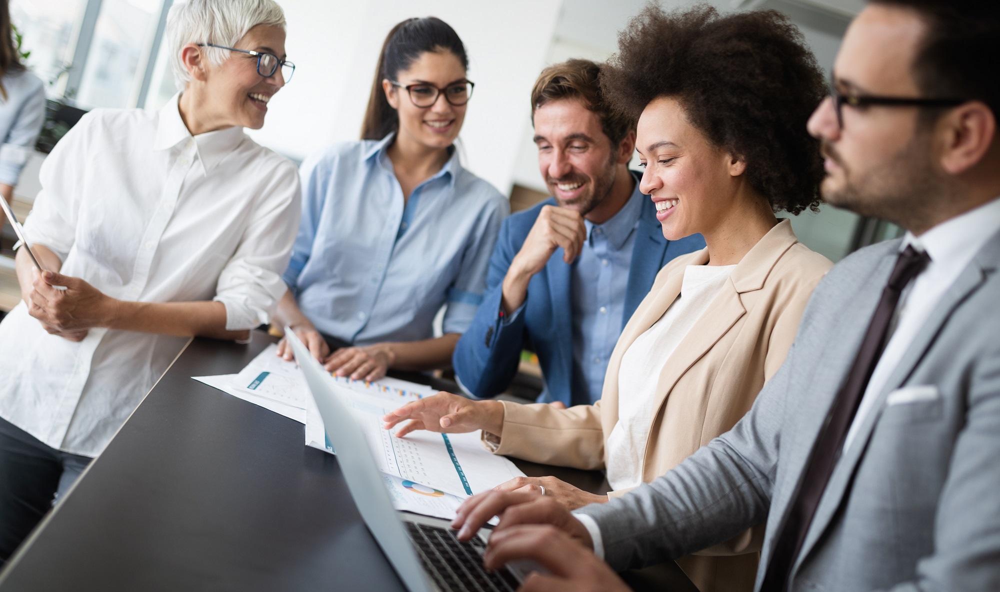 Five people standing around a desk, one person is on a laptop.  The people are looking at documents spread out on the desk in front of them.  They are smiling.
