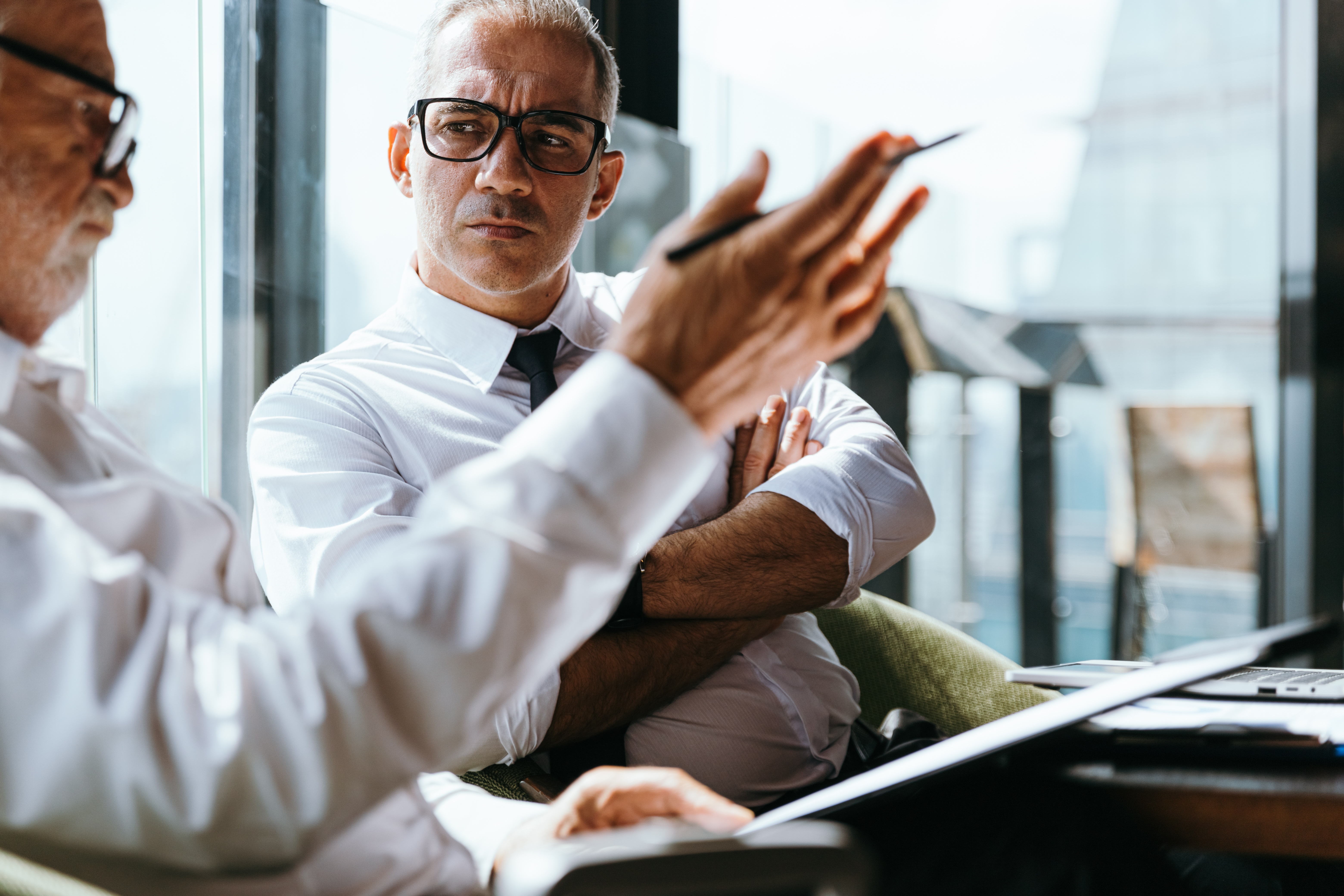Two men sat on a couch in front of a window.  One man is reviewing a document with a pen in his raised hand, the other man is sat to the right with his arms crossed.