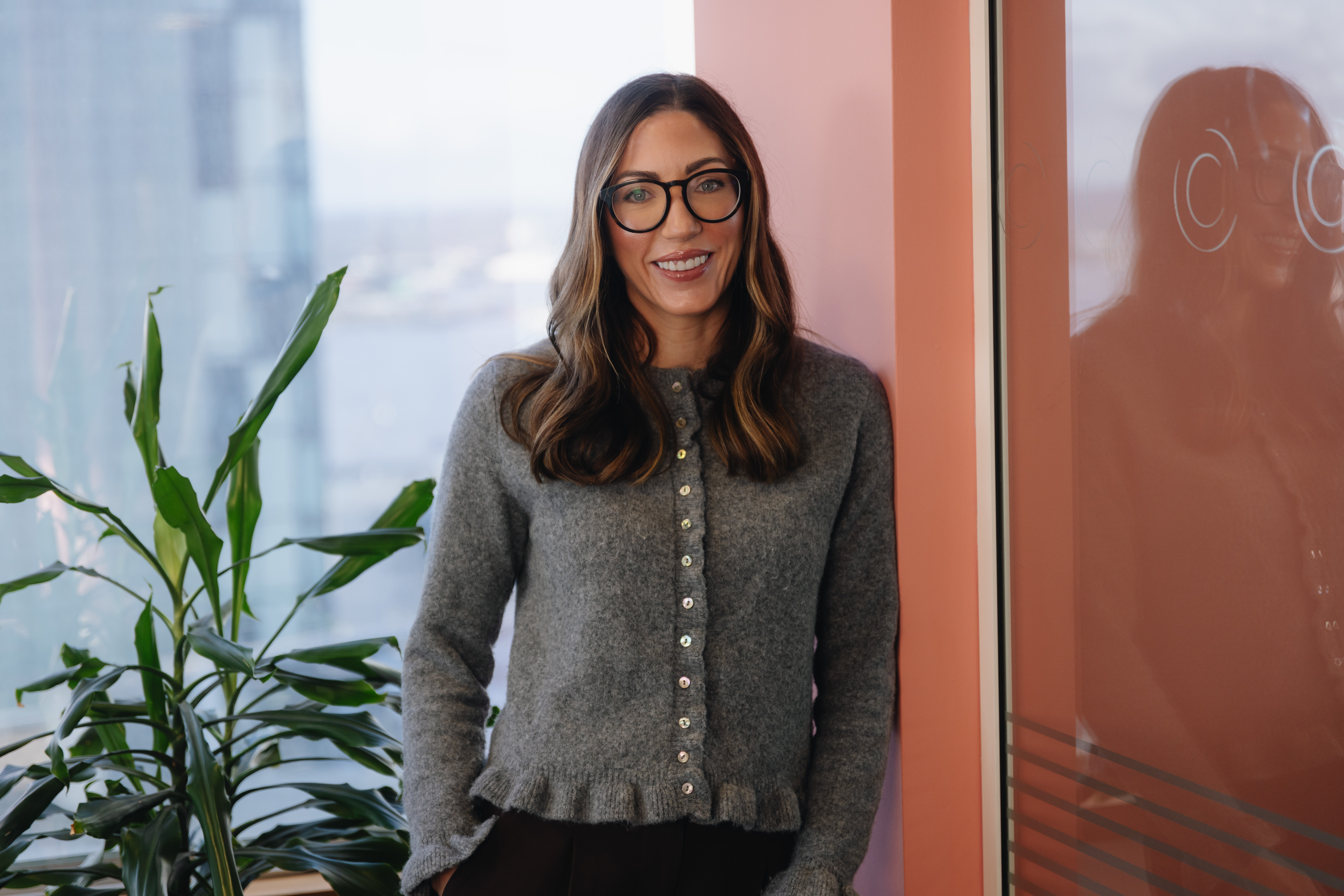 Natasha smiling leaning on a blush coloured wall to her right.  There is a window in the background with a tall green plant in front of it.