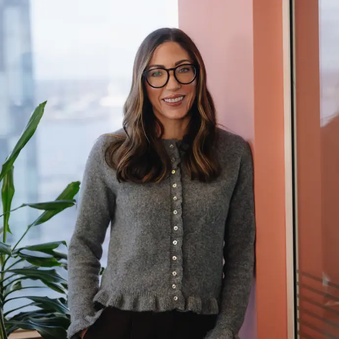 Natasha smiling leaning on a blush coloured wall to her right.  There is a window in the background with a tall green plant in front of it.