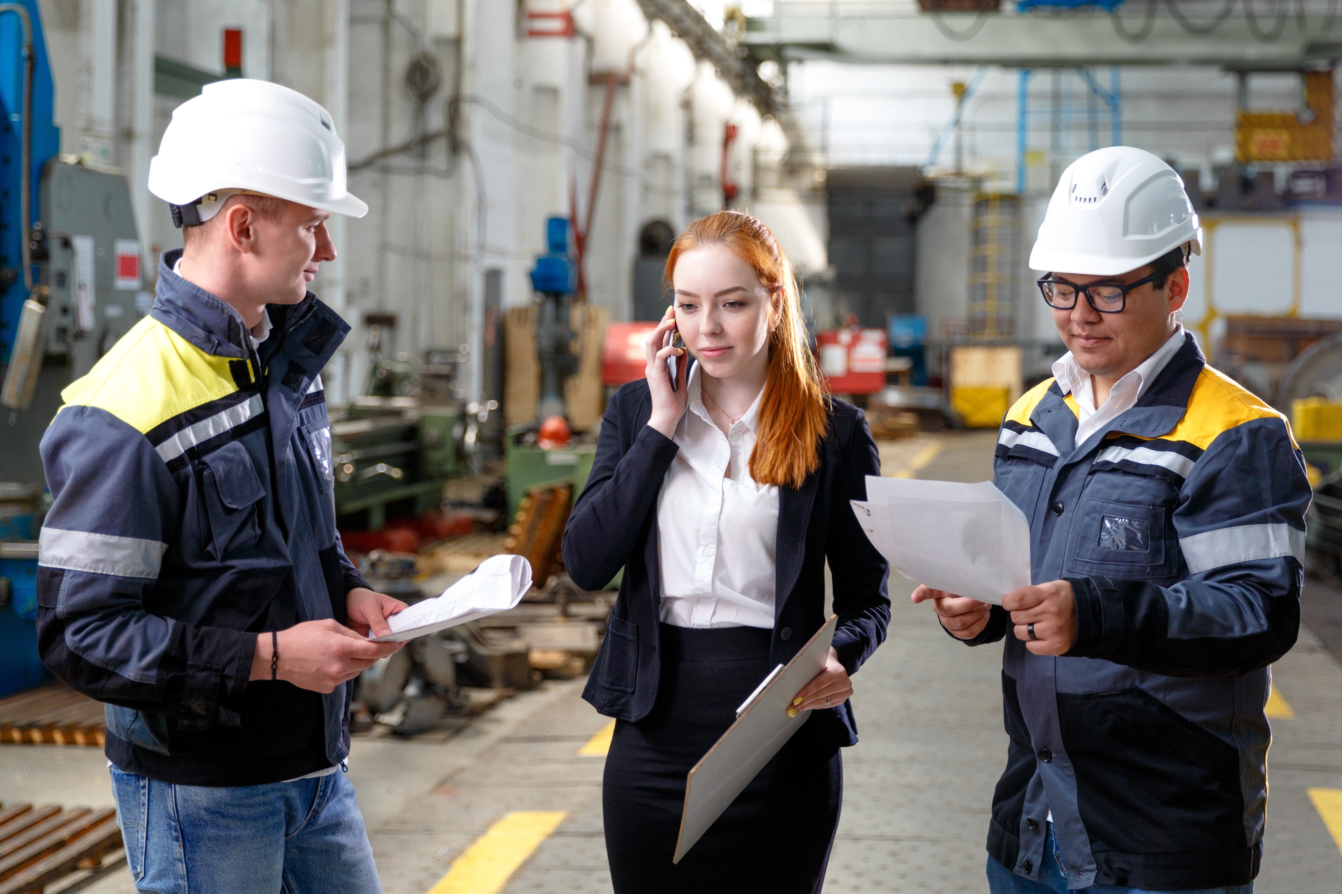A supervisor in a warehouse holding a clipboard talking on her mobile phone.  A worker holding paperwork is stood on either side of her.