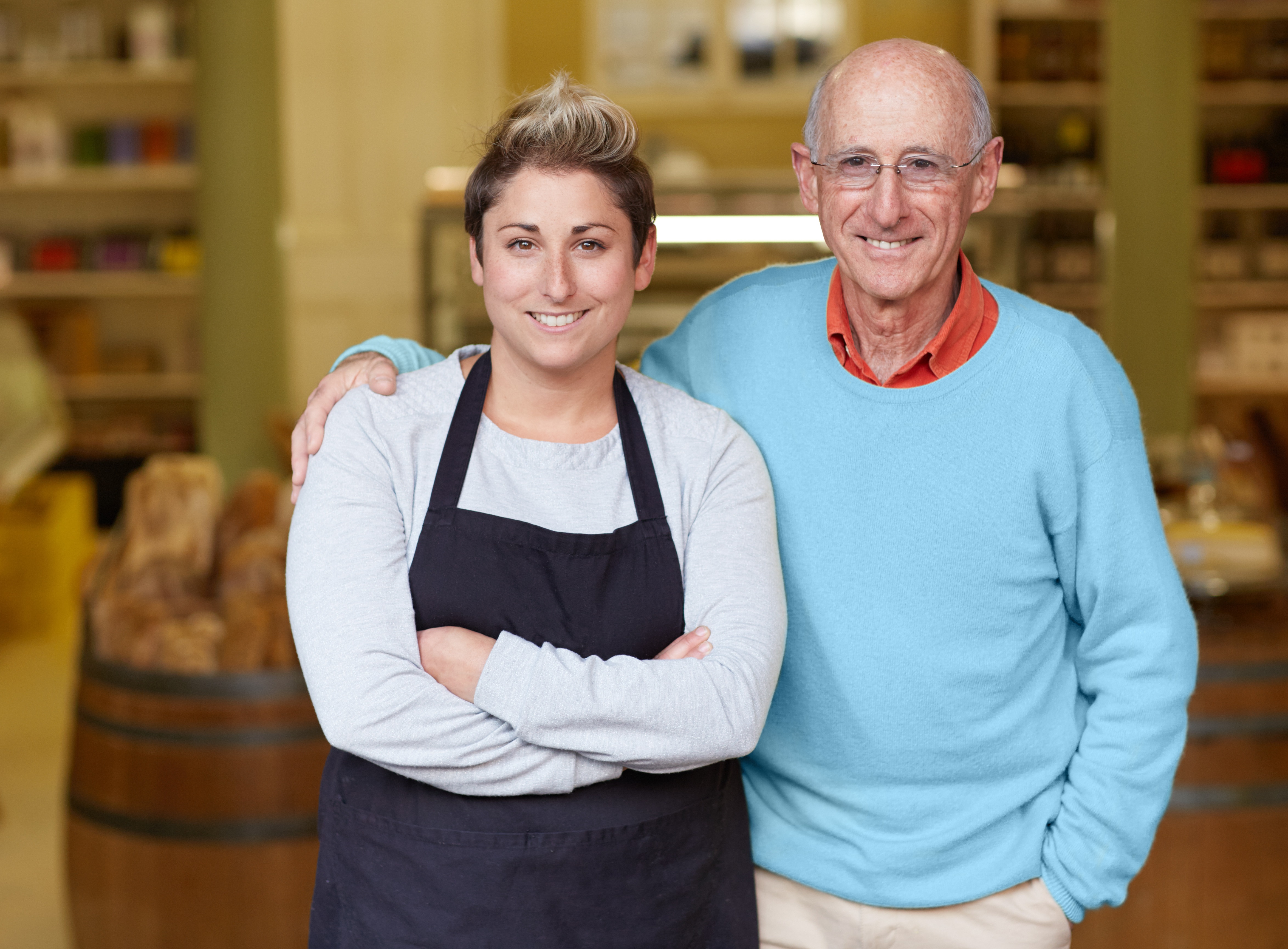 A gentleman standing next to a lady in a shop.  The lady is wearing an apron, and the gentleman has his arm around her shoulder.  There is a barrel and some shelving in the background.