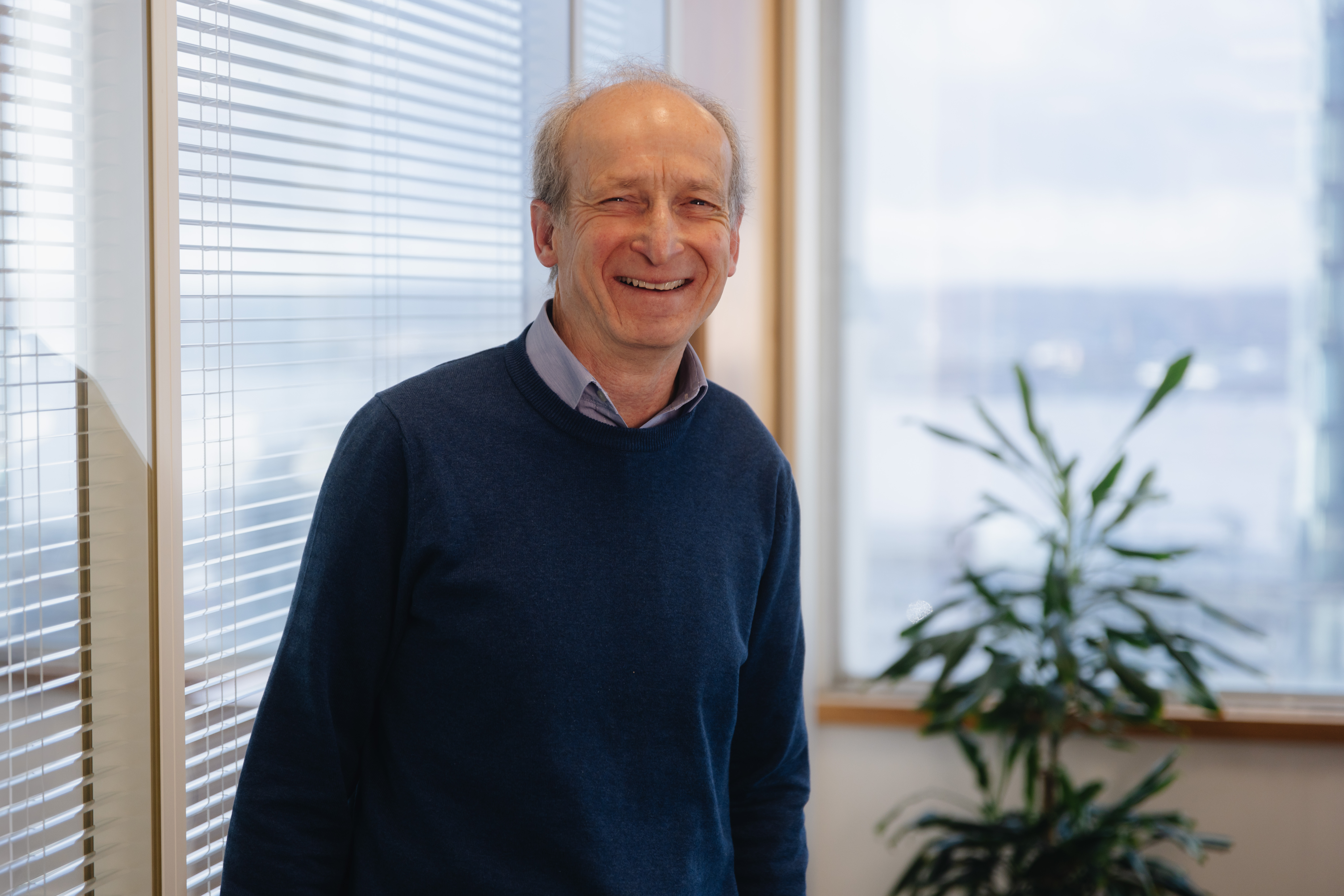 Mark smiling, leaning on a glass partition with vertical blinds.  There is a window and a tall green plant in the background to his right.