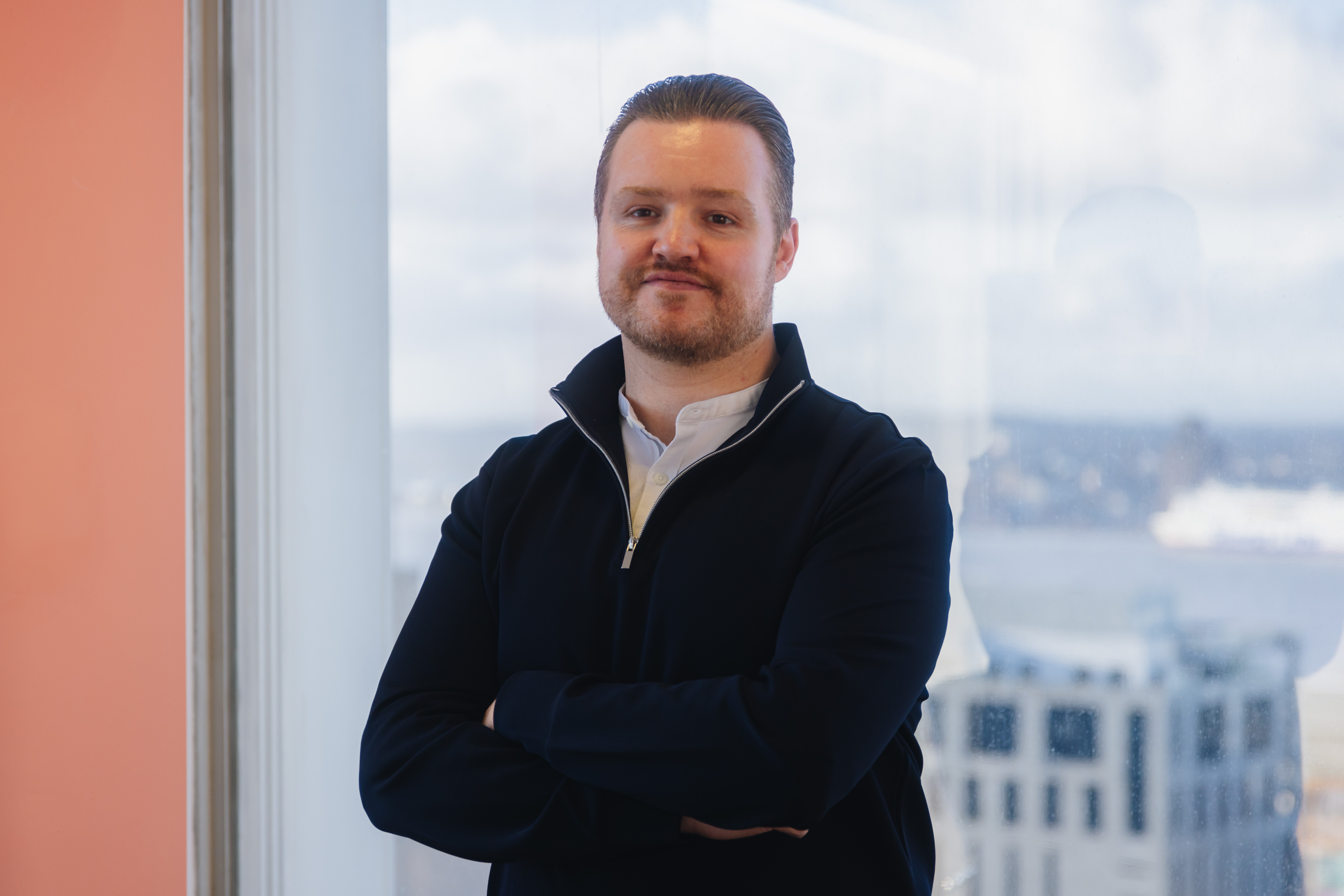 Michael stood in front of a window with his arms crossed.  He is smiling.