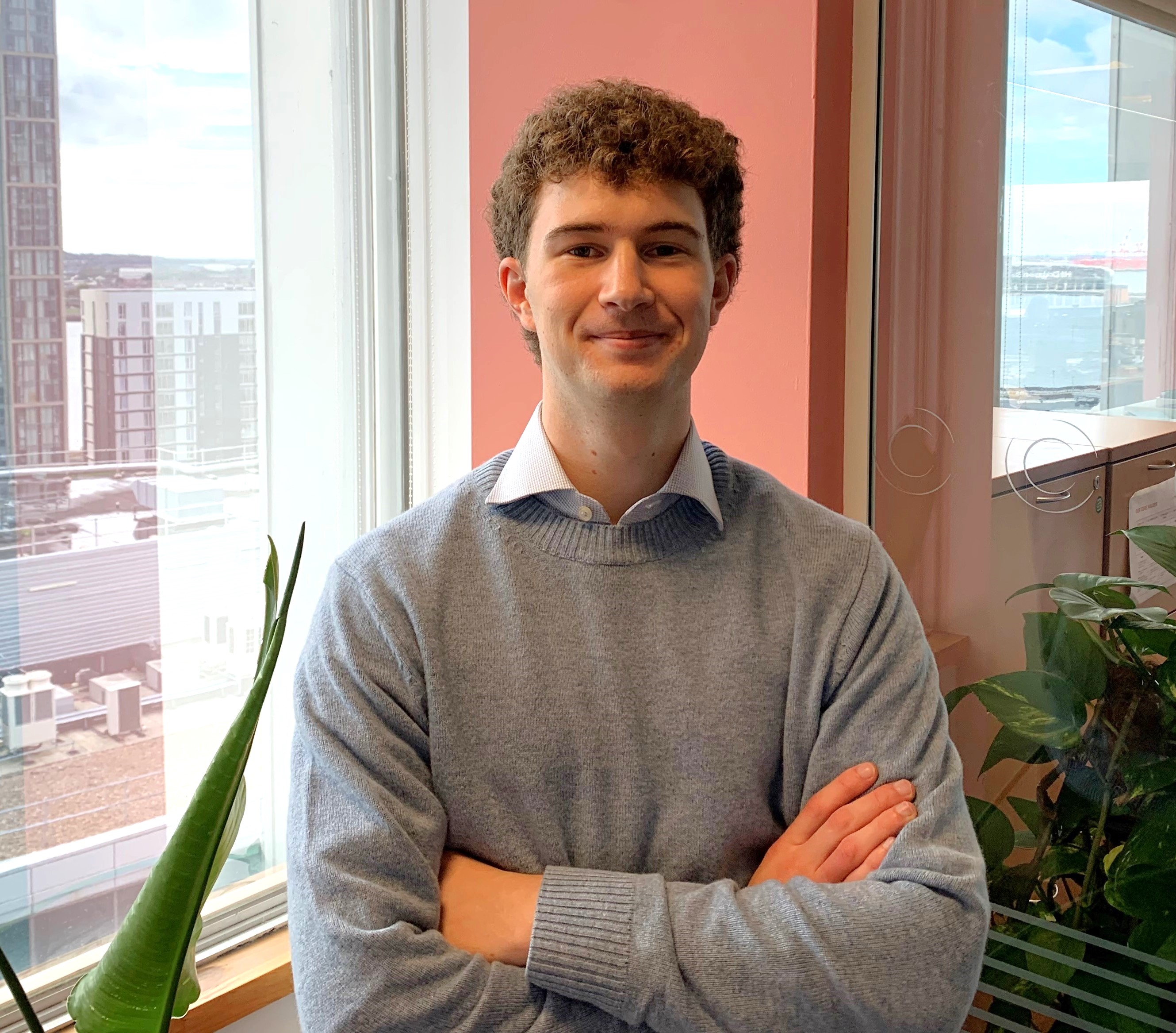 Luke stood with his arms crossed and smiling.  He is stood in front of a blush coloured pillar with a tall green plant on either side of him.