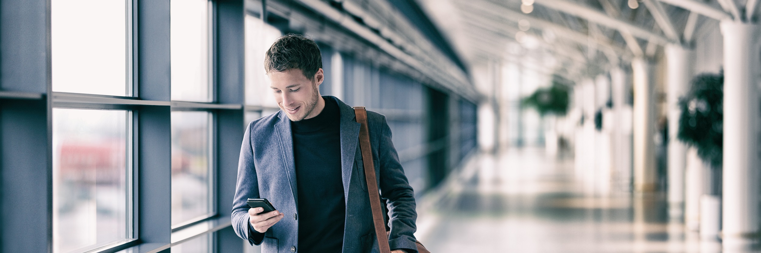 A man walking along a corridor looking at his mobile phone.  There are windows to the left and columns to the right.