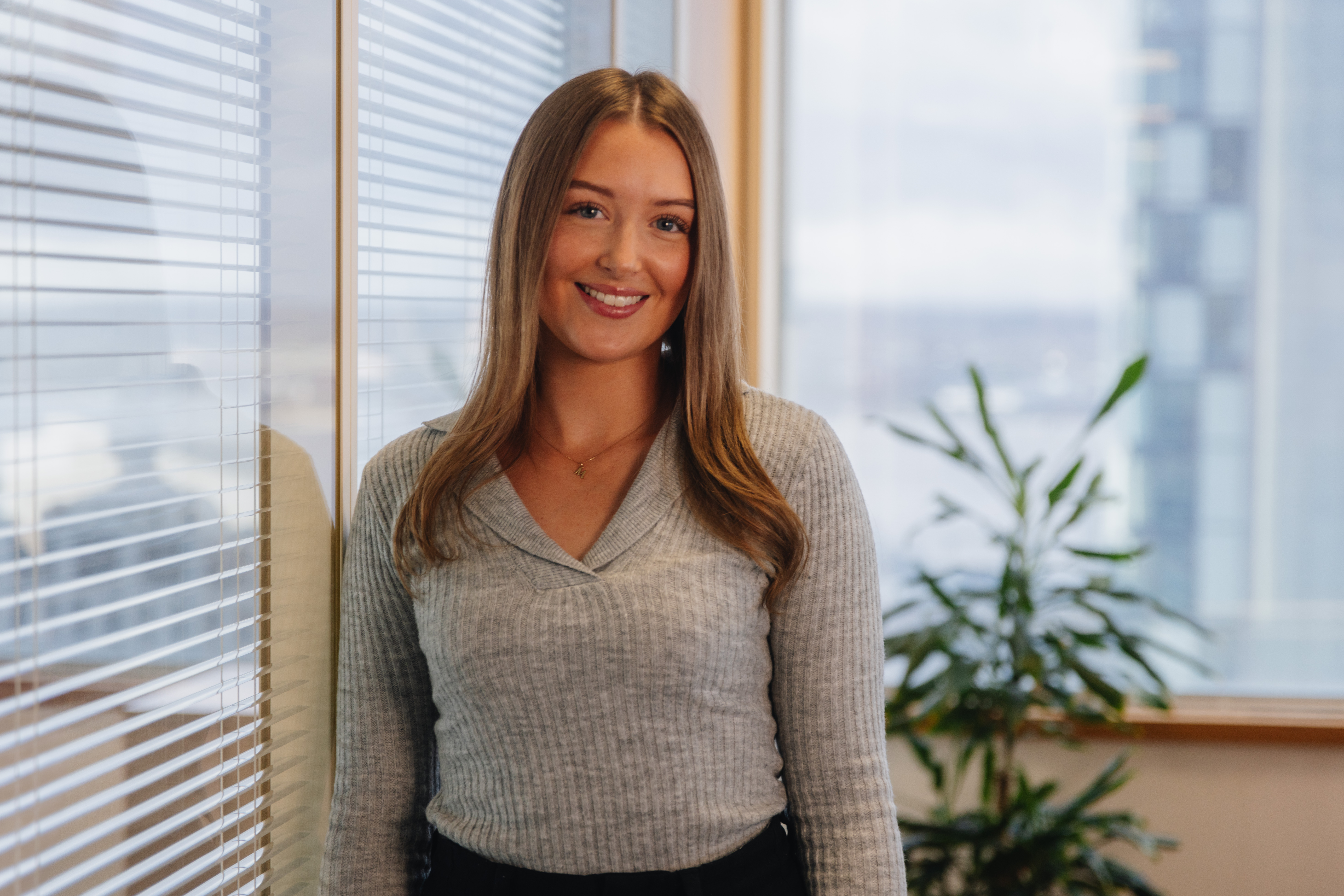 Megan standing in front of a window with a glass partition to her left.  There is a tall plant in the background.  She is smiling.