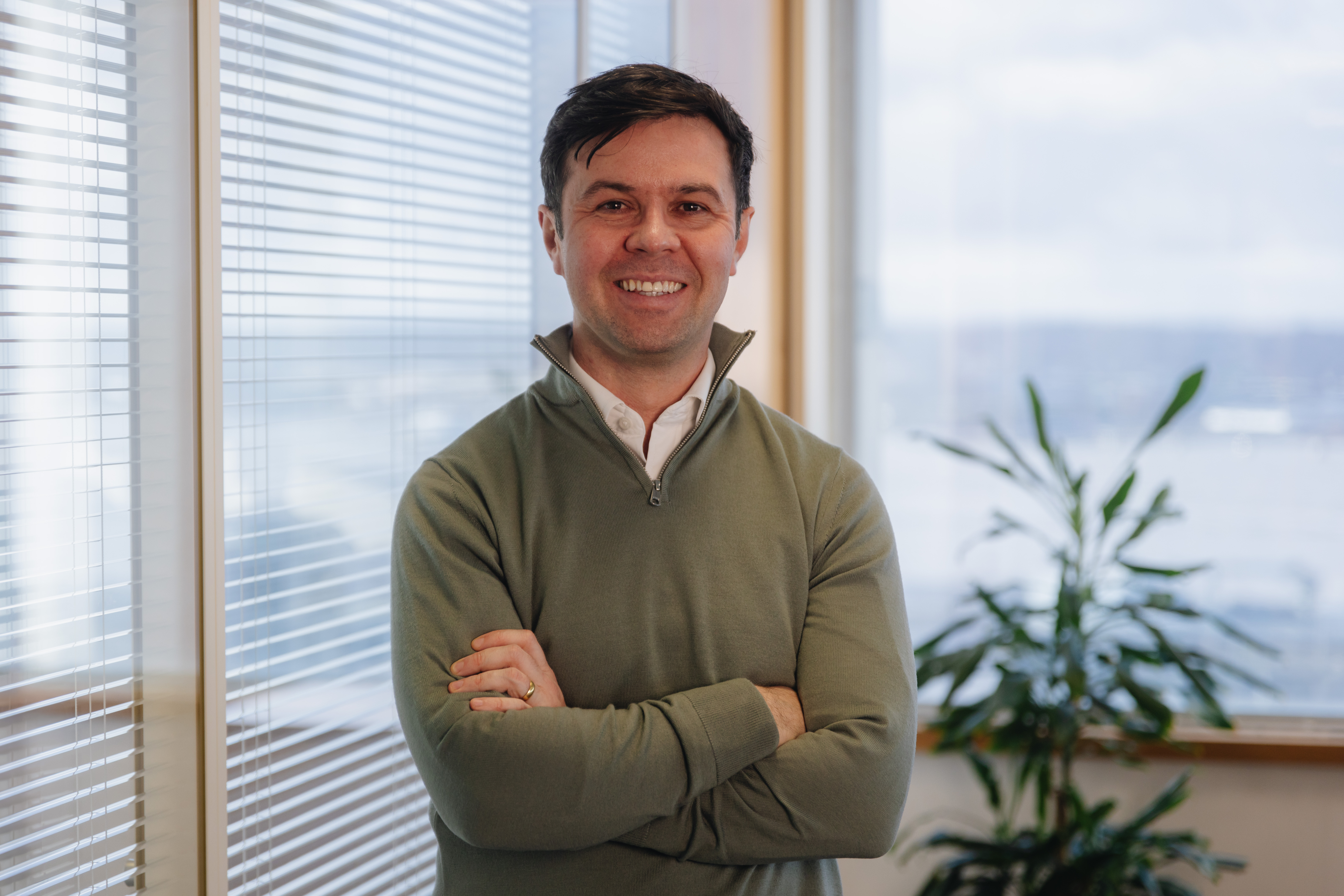 Craig standing with arms crossed and smiling next to a glass partition with vertical blinds with a window and a tall green plant in the background.