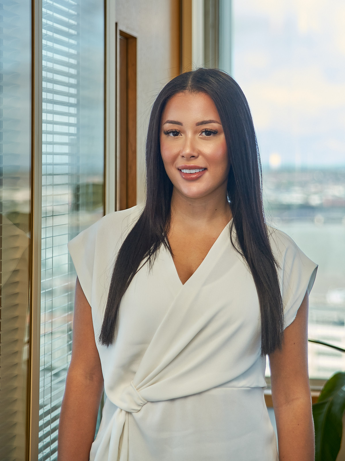 Charntai smiling stood next to a glass partition with vertical blinds.  There is a window in the background with a green plant to the right.