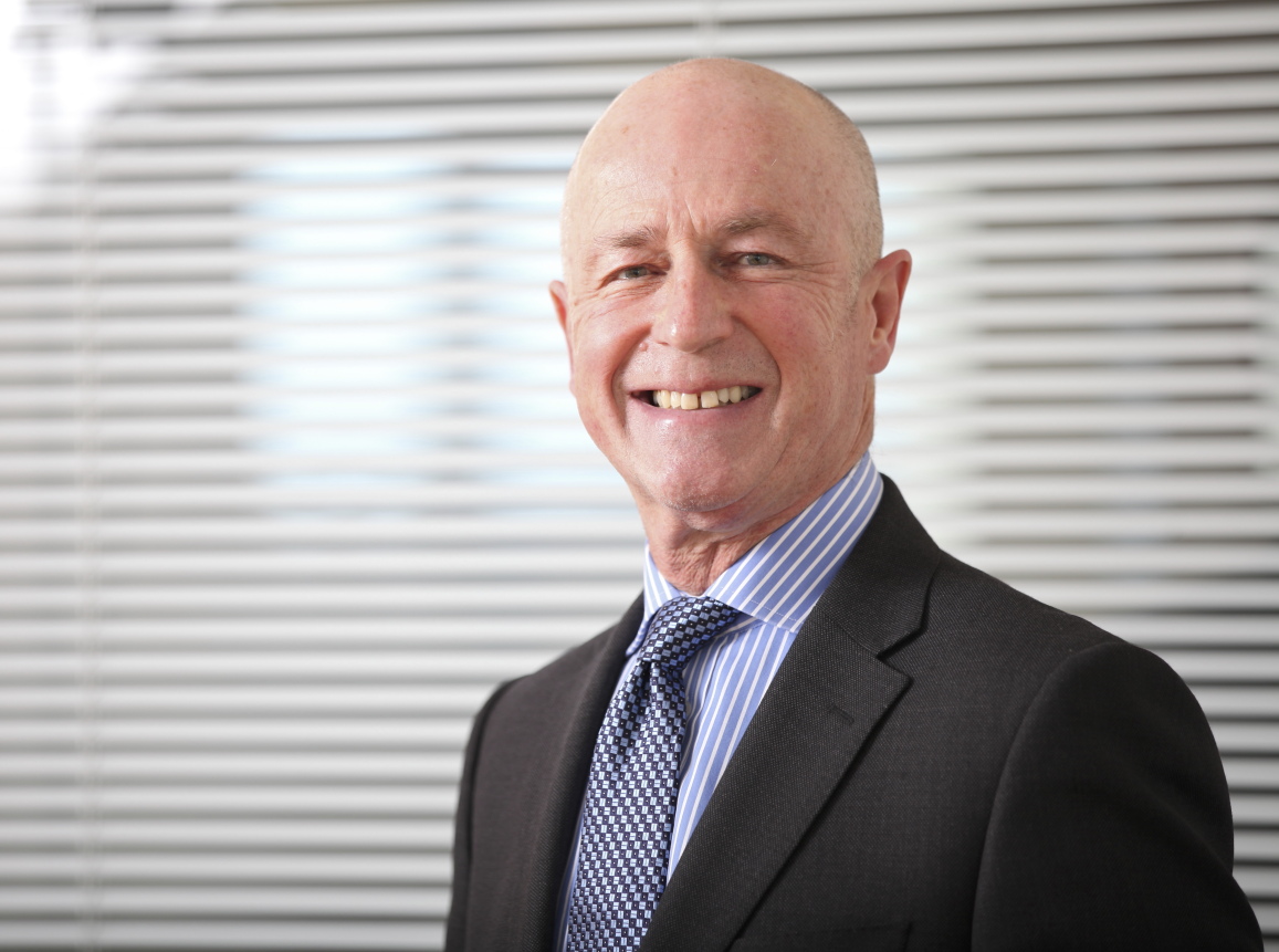 Paul smiling, stood in front of a glass partition with vertical blinds.