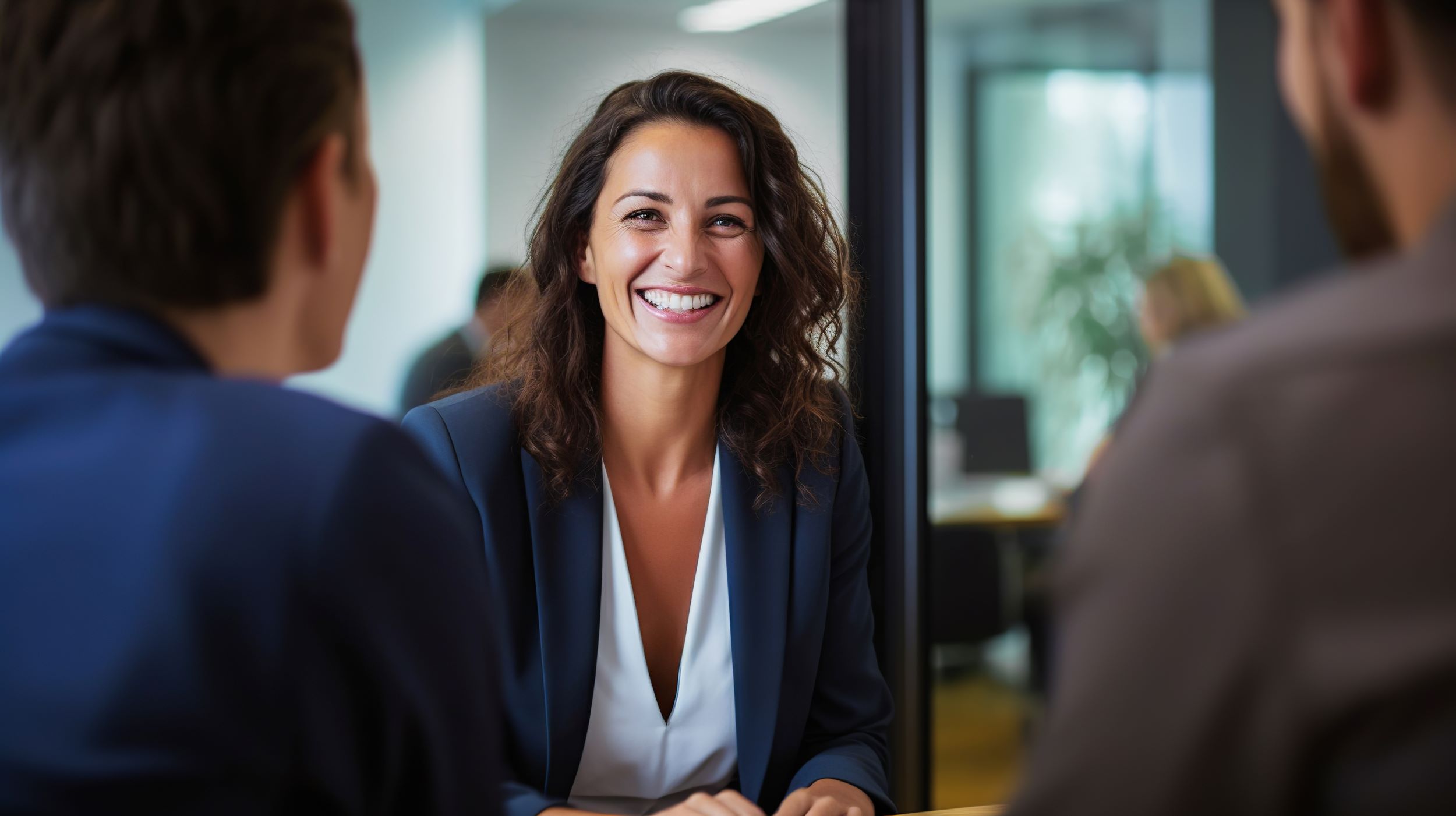 Smiling lady sat in front of a glass partition being interviewed by two people.