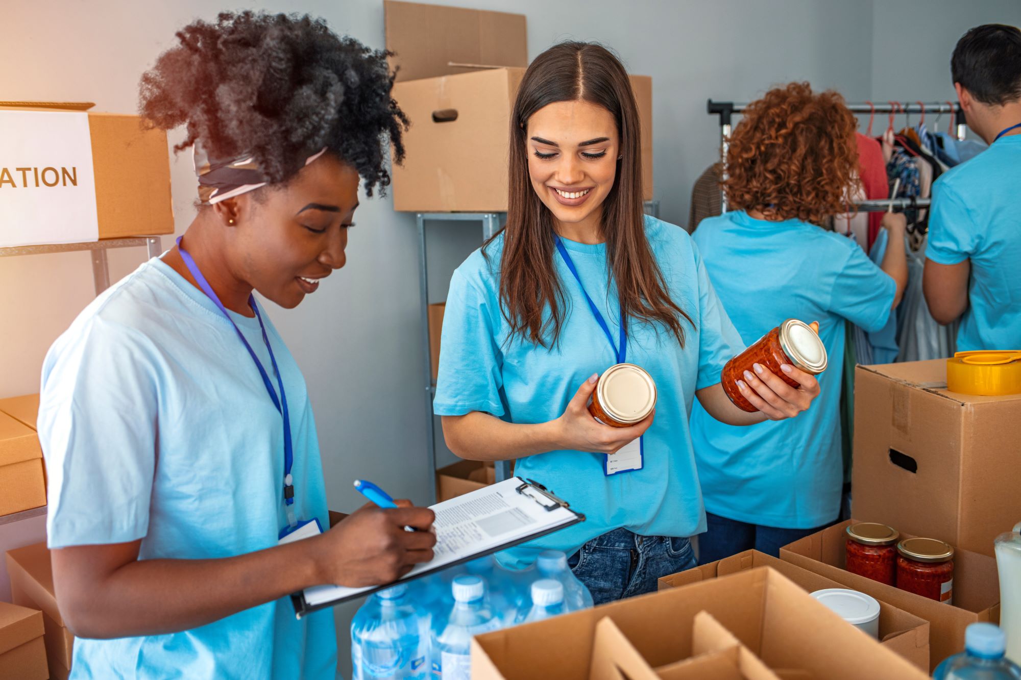 Four people sorting through donations.  The two ladies at the front are smiling, one is holding two cans and the other is writing on a clipboard.  The two in the background are looking at a clothes rail.