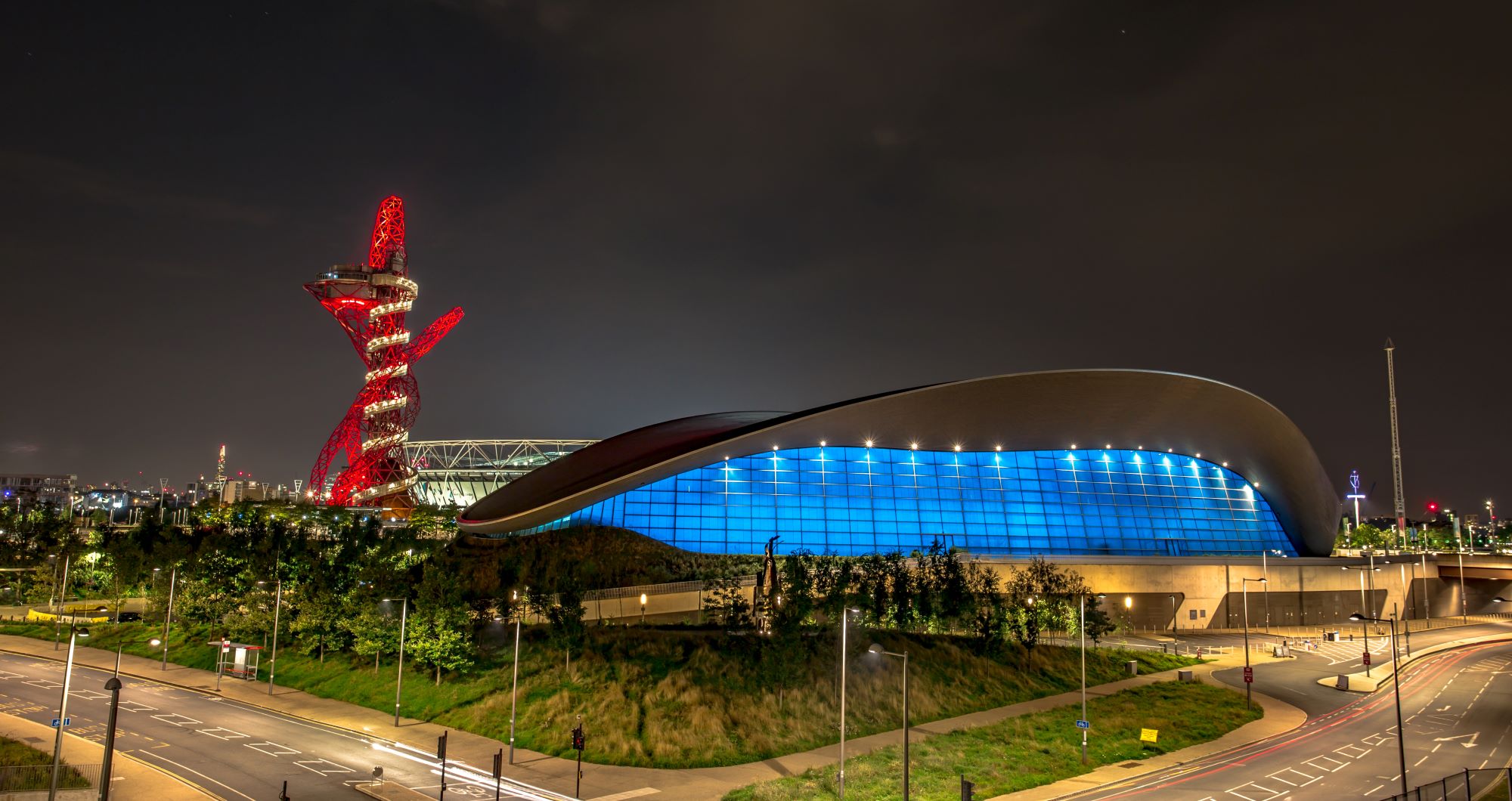 Football stadium with blue lights.  There is a tower to the left of the image lit up in red and white.  It is dark.  There is a parking lot and greenery around the stadium.