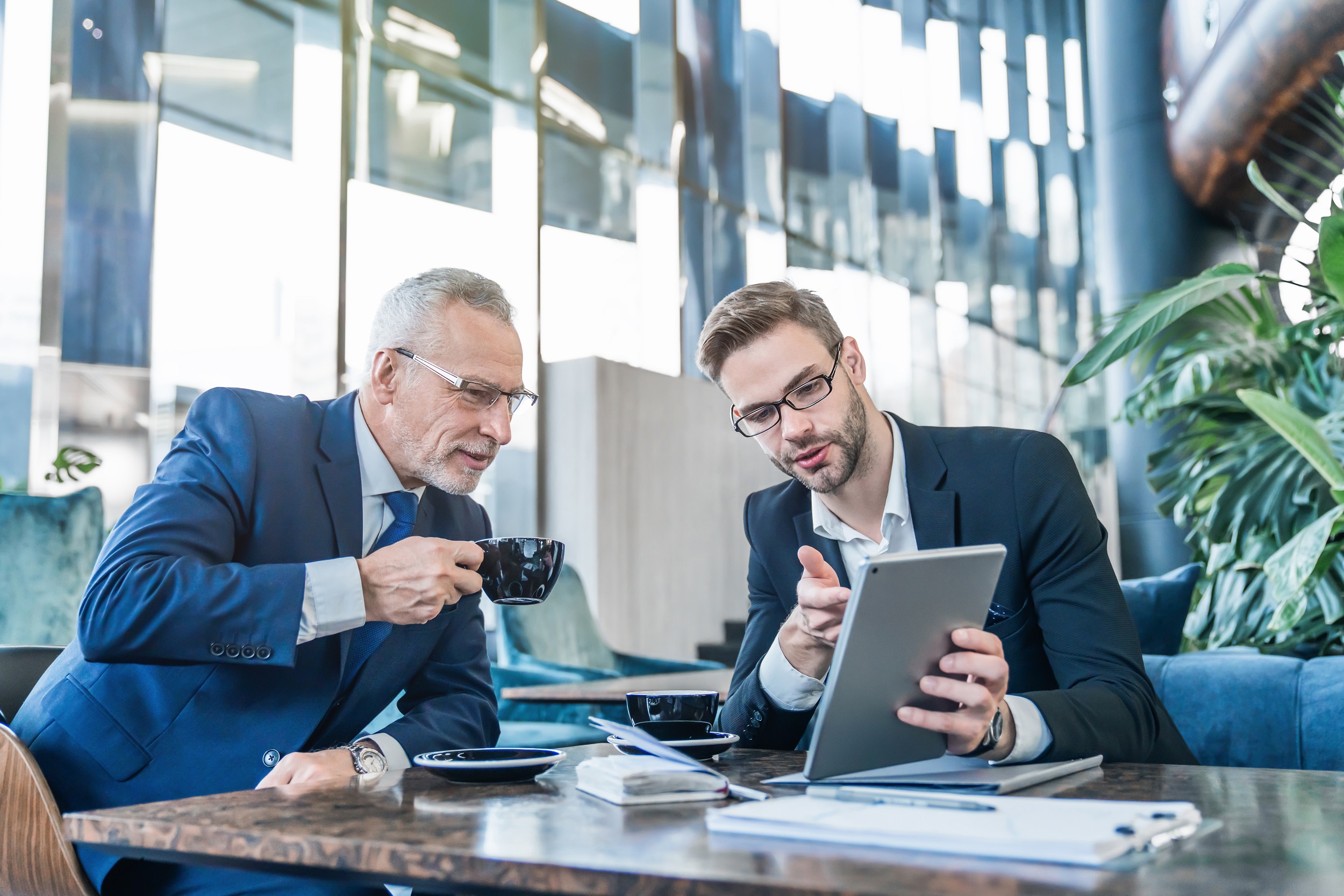 Two gentlemen sat at a table looking at a handheld tablet with documents on the table.  One man has a cup in his hand.