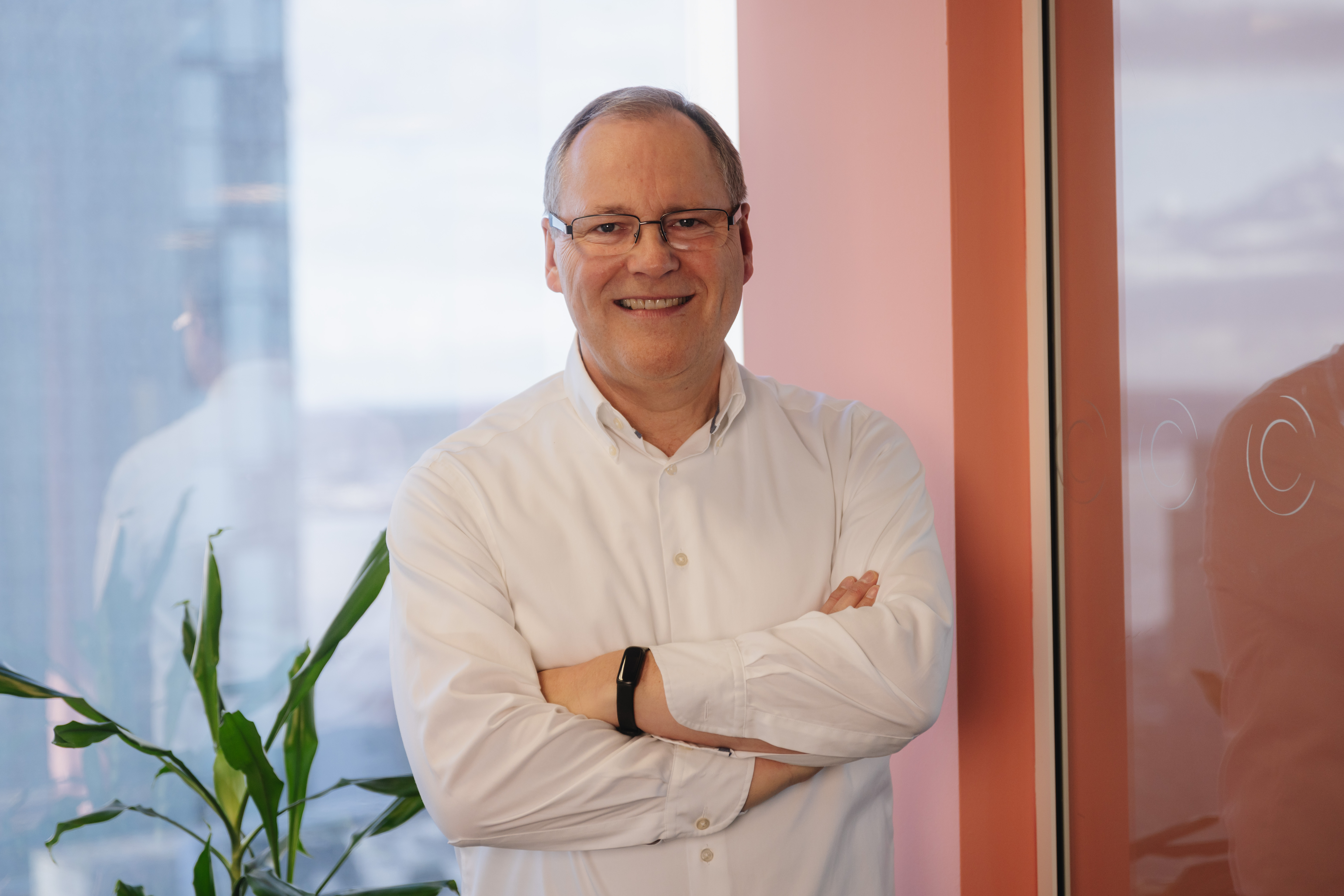Nigel stood with his arms crossed and smiling.  He is stood in front of a window with a blush coloured pillar on his right and a tall green plant on his left.