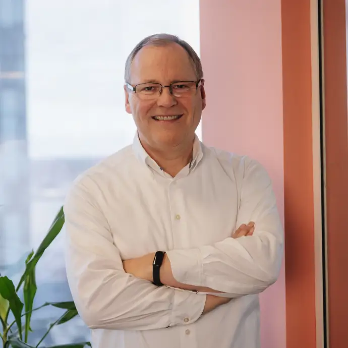 Nigel stood with his arms crossed and smiling.  He is stood in front of a window with a blush coloured pillar on his right and a tall green plant on his left.